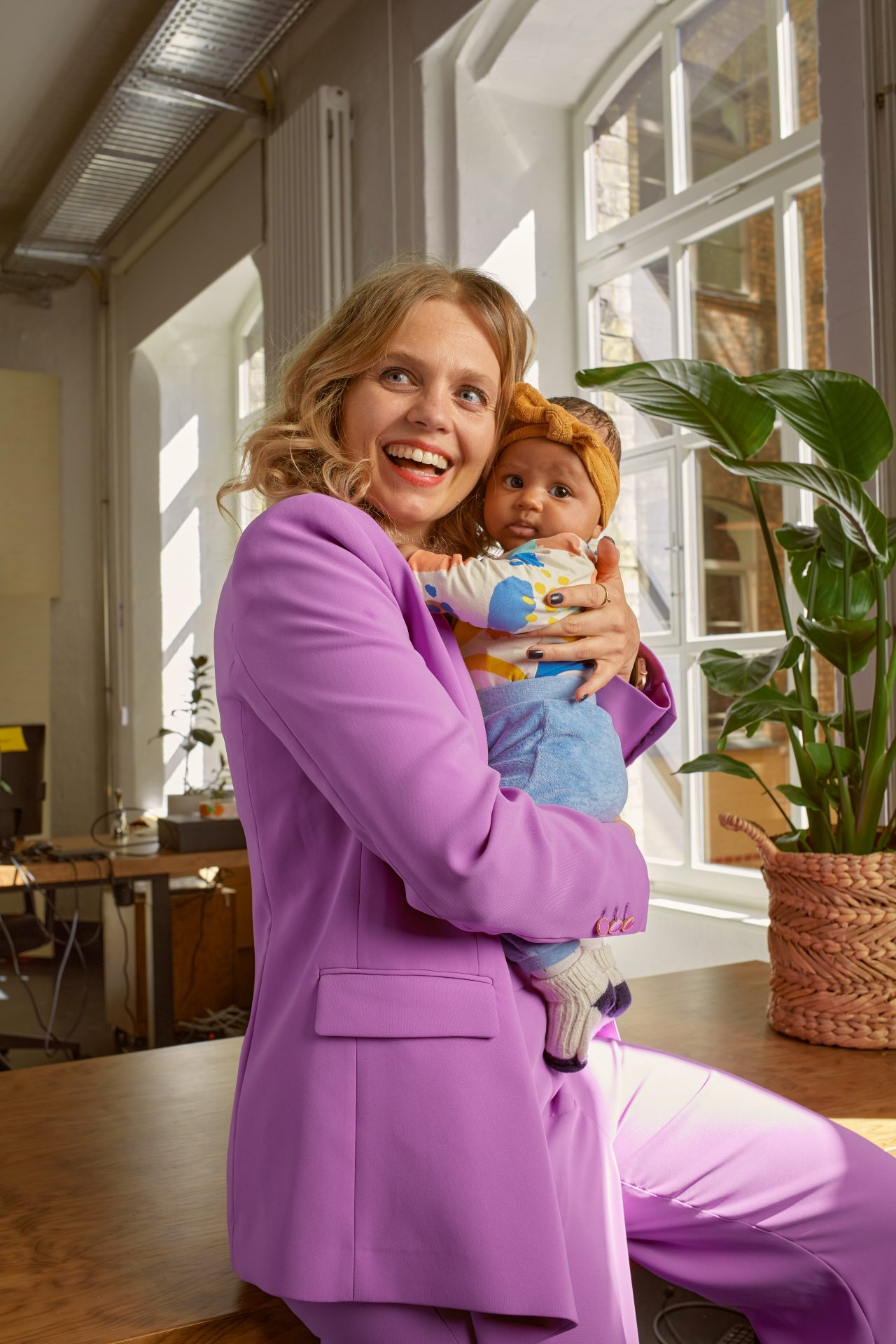 Eine blonde Frau in pinken Anzug lächelt und hält ein Baby n der Hand.