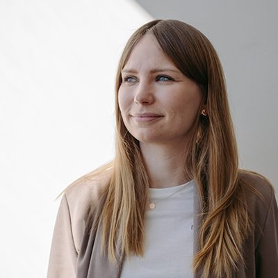 Frau mit hellbraunen Haaren steht vor einer weißen Wand und ihr Gesicht ist halb im Schatten. Sie trägt einen weißen T-shirt und beigen Blazer  und ihr Gesicht ist nach links gerichtet.