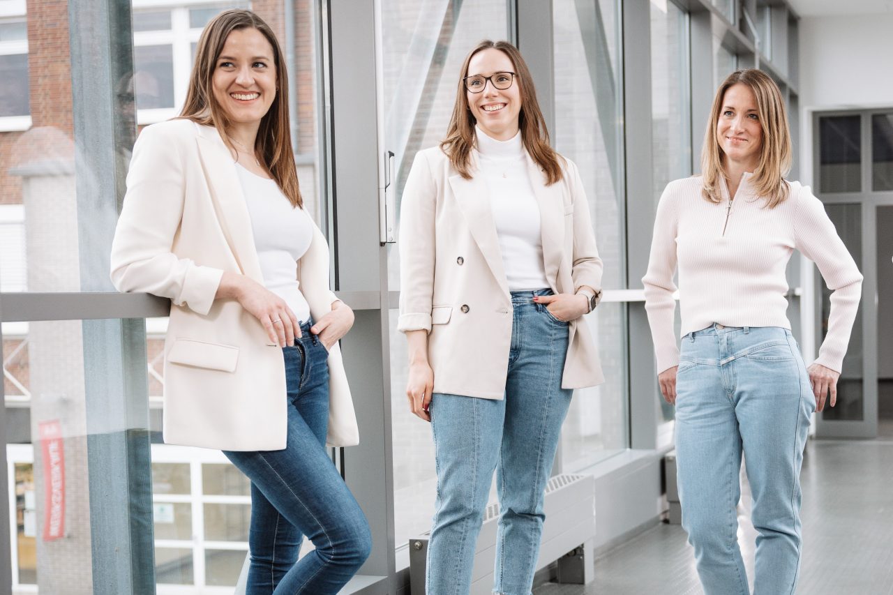 Drei Frauen, Recruiterinnen bei Melitta, stehen in einem Lichtgang zwischen zwei Gebäuden und lächeln in die Kamera