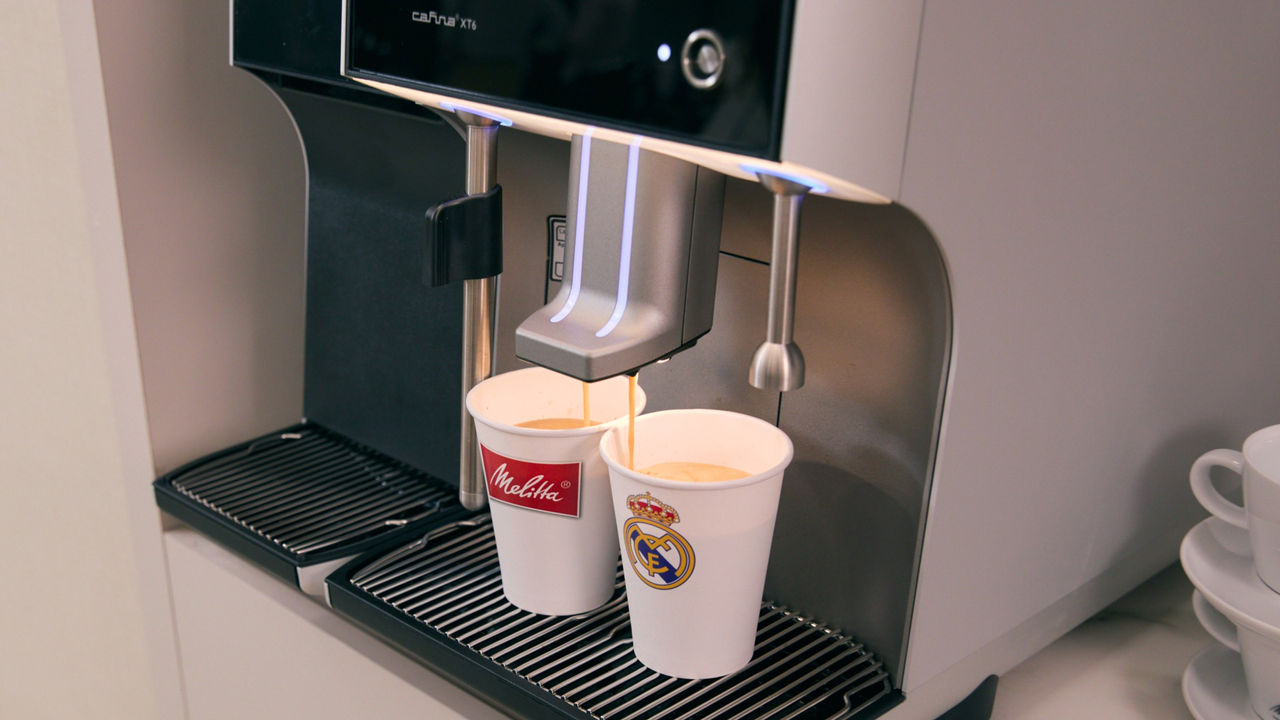Eine Melitta-Kaffeemaschine (Modell XT6) brüht gleichzeitig Kaffee in zwei Becher - einen mit Melitta-Logo und einen mit Real Madrid-Wappen. Frisch gebrühter Kaffee fließt in beide Tassen.