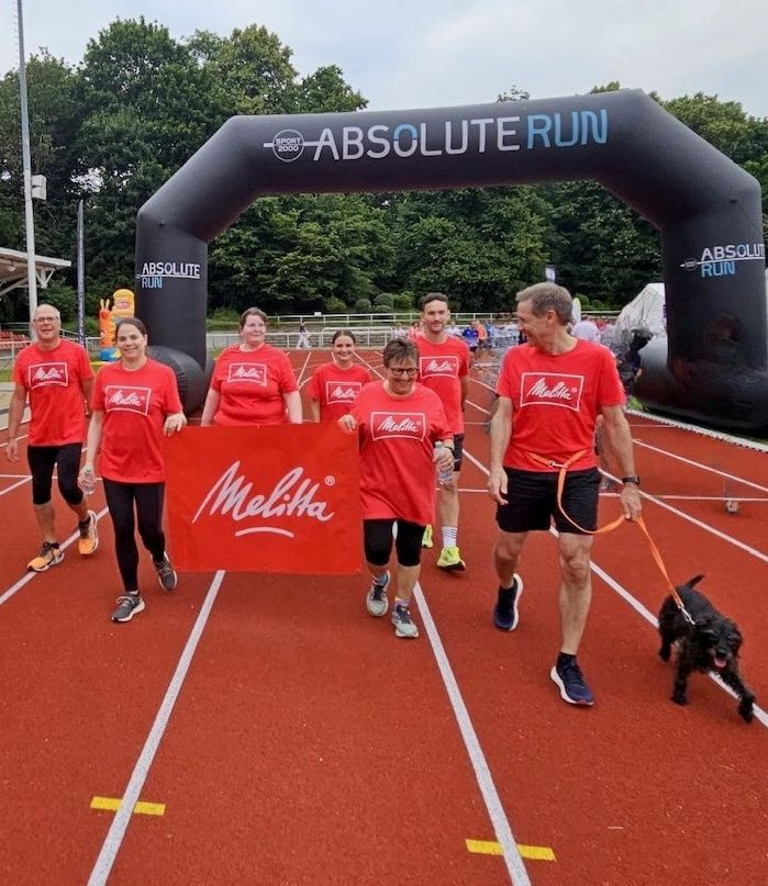Eine Gruppe von Menschen in roten Melitta-T-Shirts und mit Hund geht gemeinsam unter einem Startbogen über die Laufbahn und hält ein Melitta-Banner. [KI-bearbeitet]]