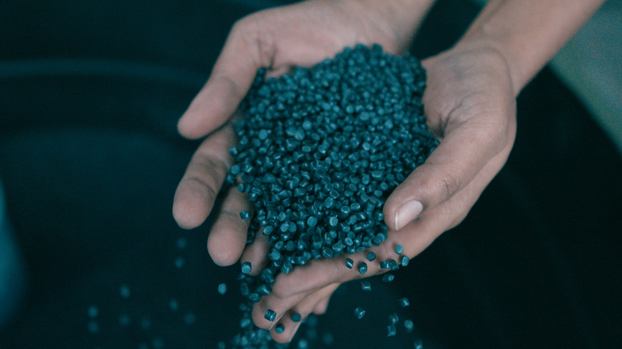 Zwei Hände halten eine Portion türkisfarbener, recycelter Kunststoffpellets, weitere Pellets rieseln herab.