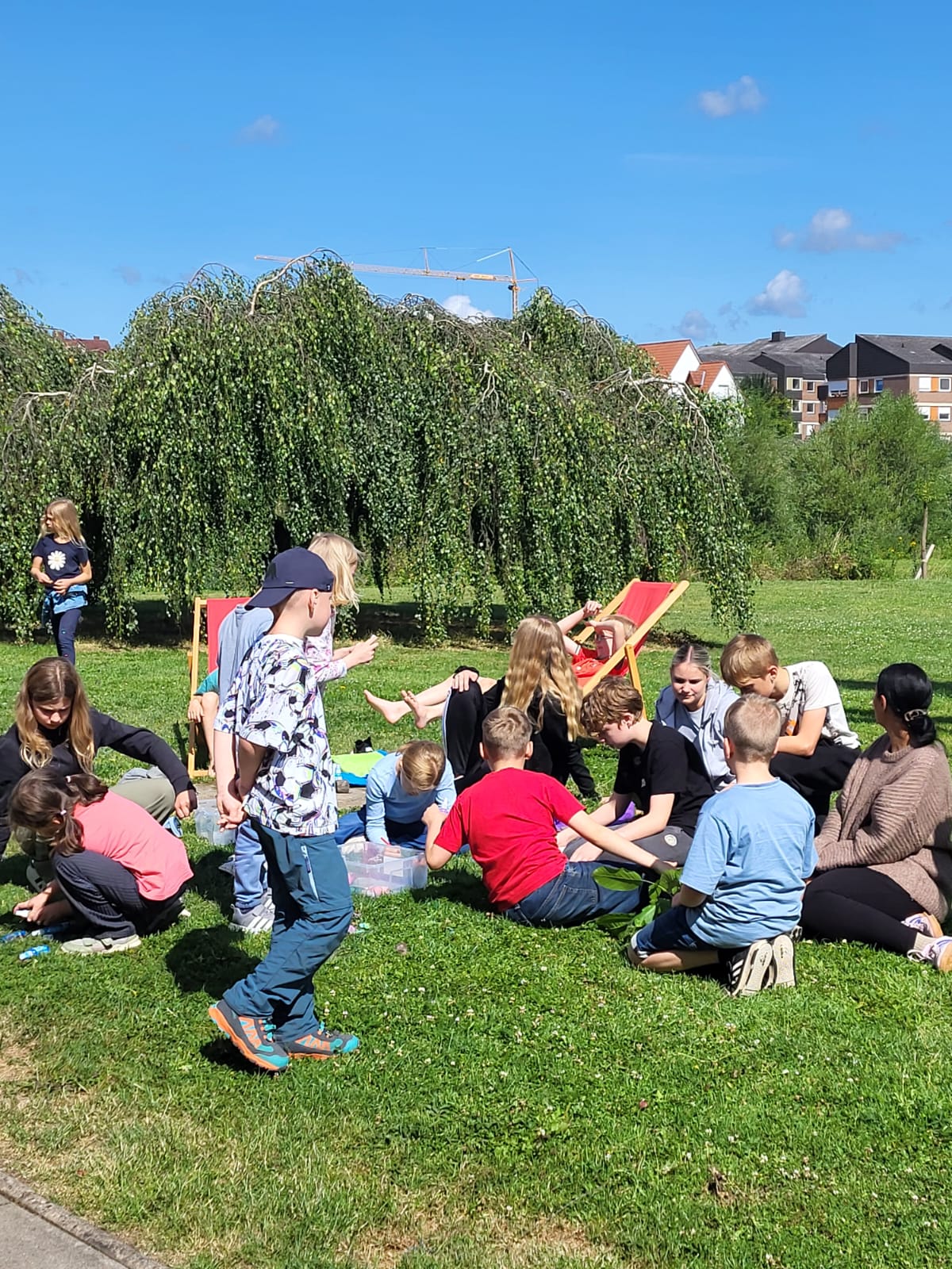 Gruppe von Kindern und Betreuerinnen sitzt und kniet auf einer Wiese vor einer großen Trauerweide, im Hintergrund Wohnhäuser und ein Baukran unter blauem Himmel 