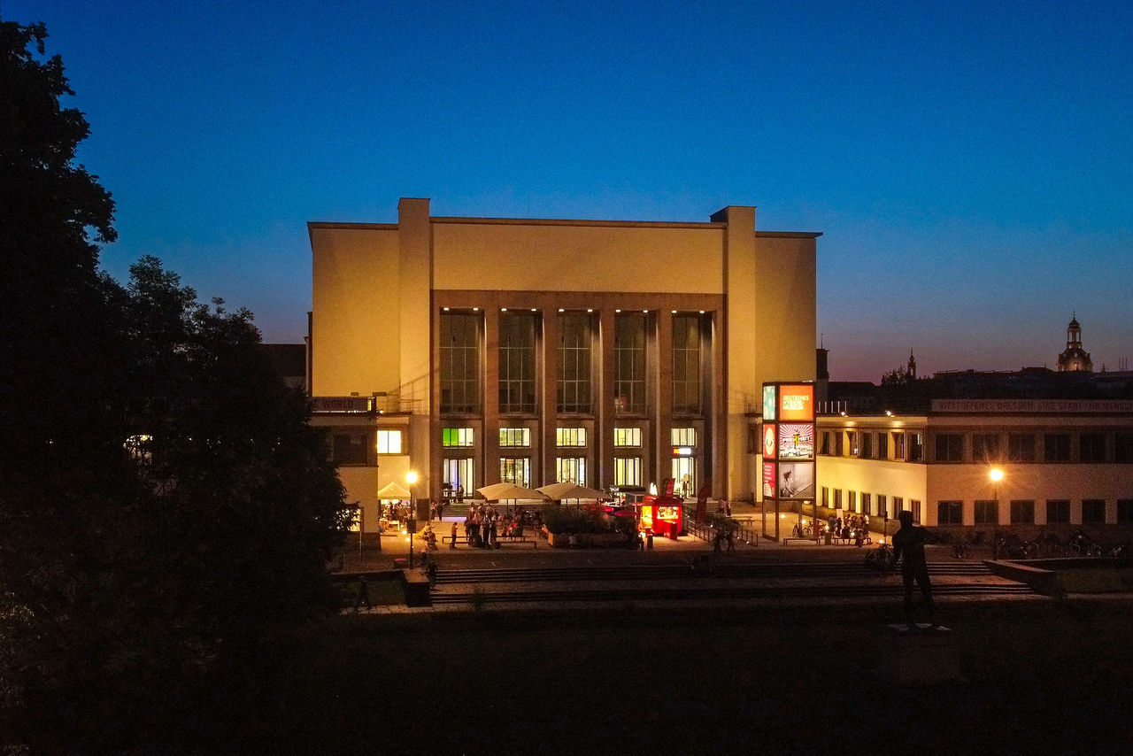 Das Deutsche Hygiene-Museum Dresden bei Dämmerung, die beleuchtete Fassade und der Vorplatz sind sichtbar, Menschen versammeln sich vor dem Gebäude.