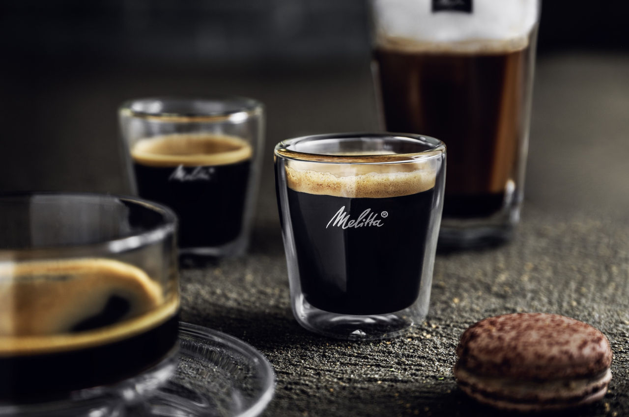 Double-walled espresso glass with crema and Melitta lettering on a dark stone background. Behind it are more glasses of coffee, on the right a macaron