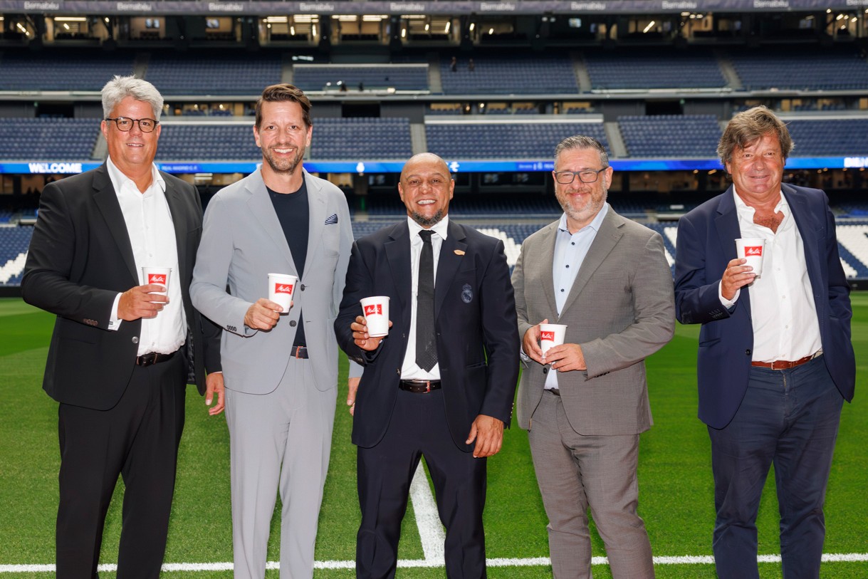Fünf Männer in Business-Kleidung stehen auf dem Rasen des Bernabéu-Stadions und halten Melitta-Kaffeebecher. Im Hintergrund sind die leeren Tribünen des Stadions zu sehen.