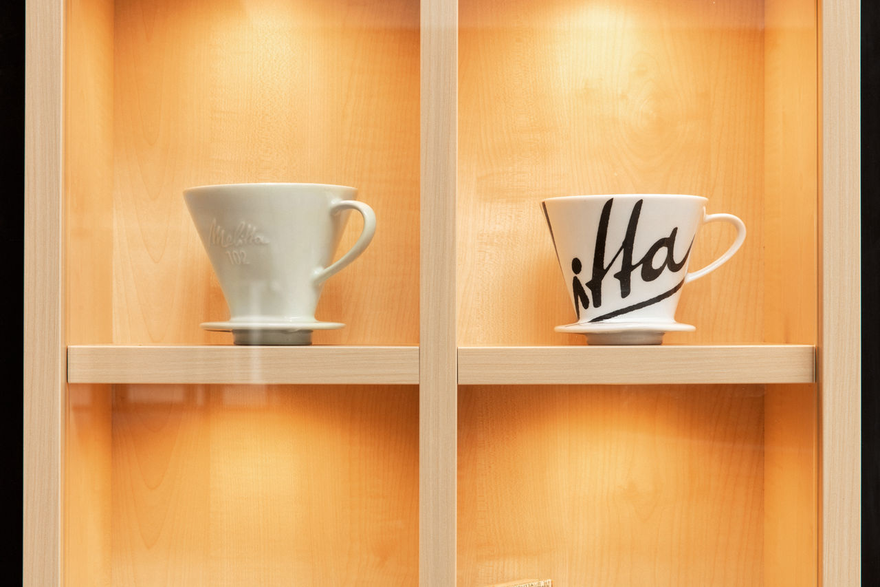Illuminated wooden display case with two white porcelain coffee filters next to each other: on the left a filter with a small Melitta 102 lettering, on the right a filter with an oversized, decorative Melitta lettering cut-out "itta".