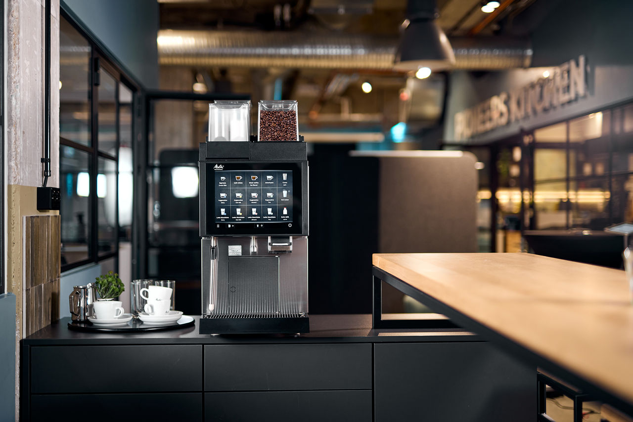 Front view of a Melitta Cafina CT8plus standing on a counter in a modern café or dining room environment with warm light and wooden elements.