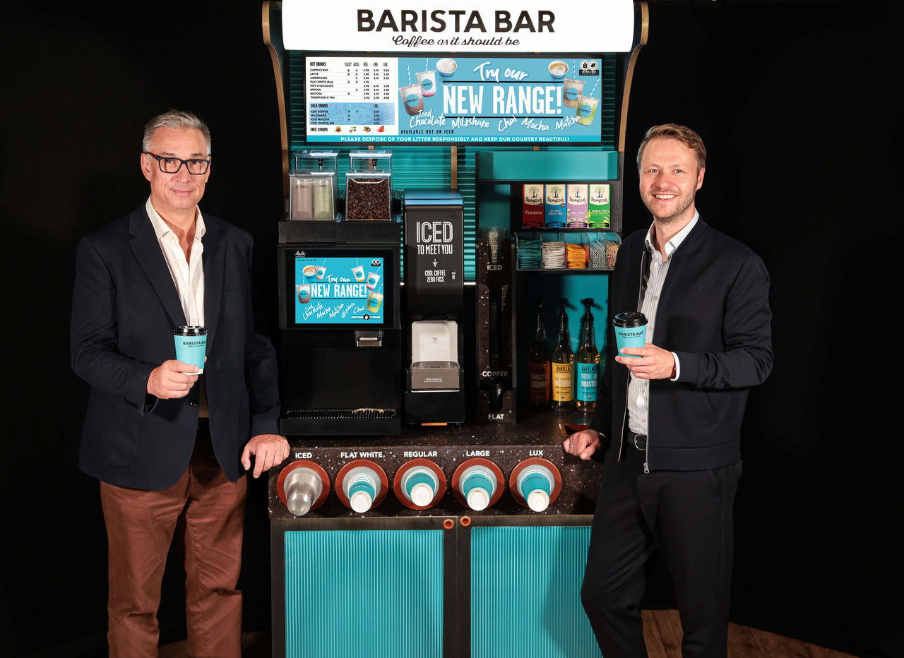 Two men in suits stand next to each other in front of a turquoise barista bar kiosk with the inscription "NEW RANGE" and each hold a coffee mug in their hands.