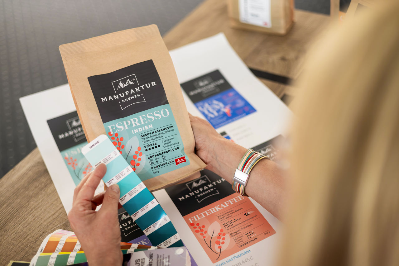 Eine Frau vergleicht einen türkisfarbenen Pantone-Farbfächer mit dem Etikett eines Kaffeebeutels der Melitta Manufaktur Bremen, Sorte Espresso Indien. Weitere Verpackungsentwürfe liegen auf dem Tisch 