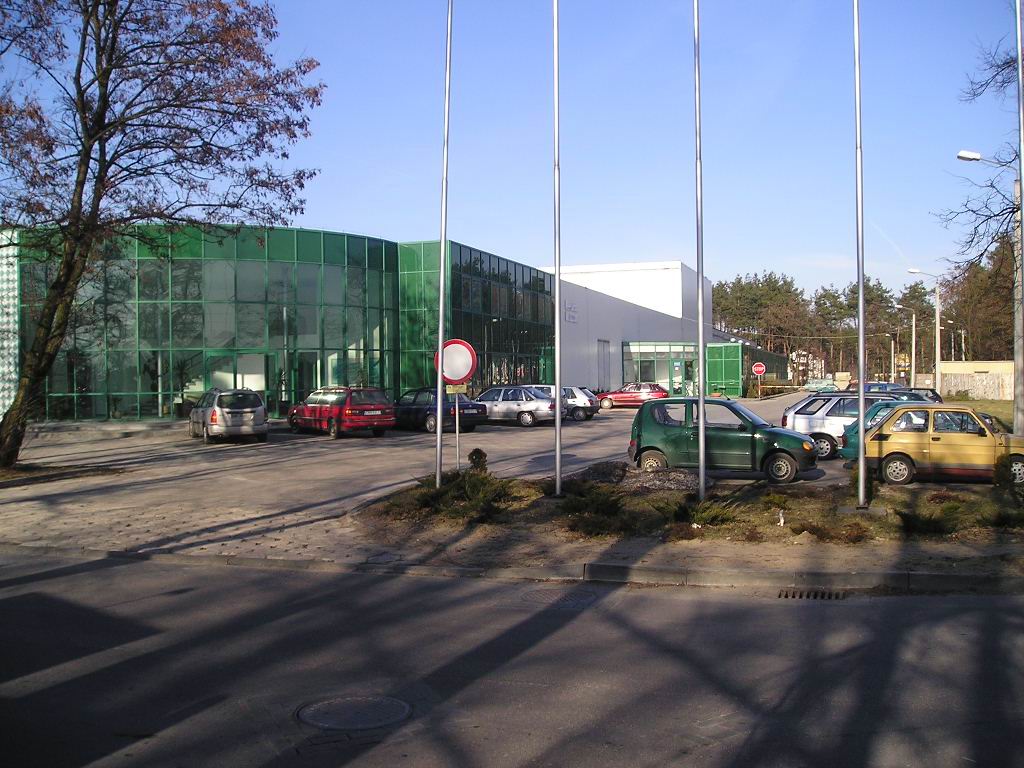 Firmengebäude mit Glasfassade, umgeben von Grünflächen, Parkplätzen und Bäumen, bei Tageslicht. 