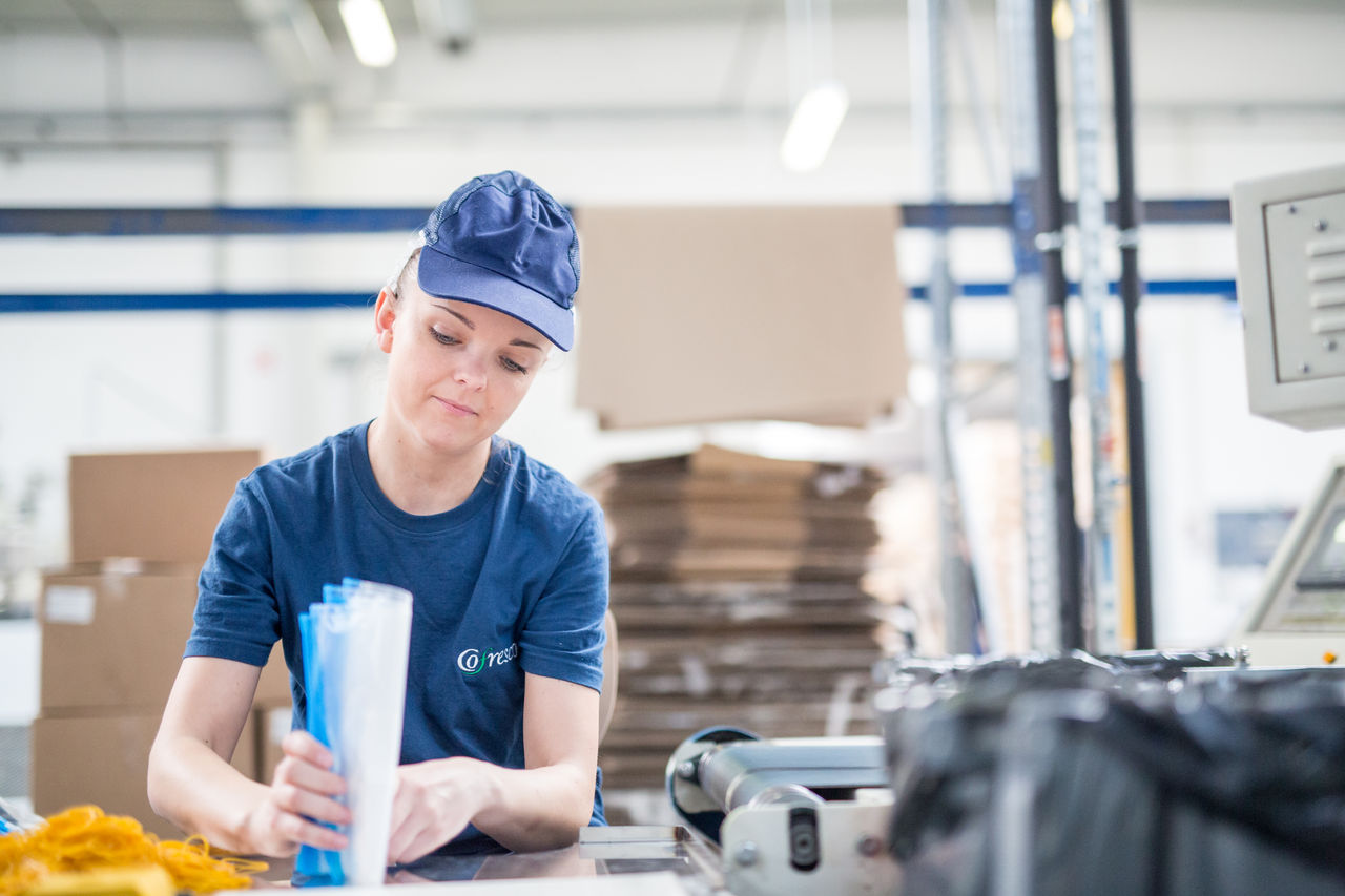 Person an einem Arbeitsplatz in einer Produktionshalle sortiert oder verpackt Folienbeutel, im Hintergrund Kartons und Maschinen